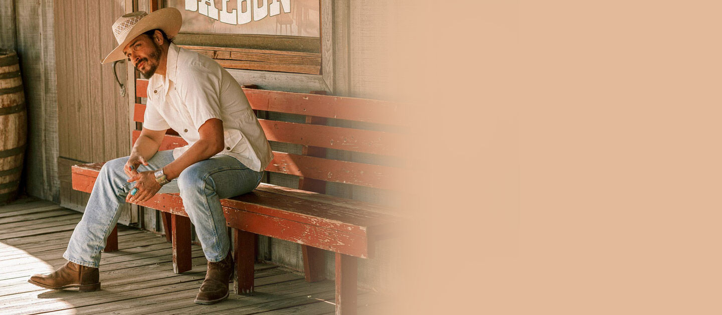 A man sitting on a bench, wearing the Bowie 11” Pull-On Western Boot in oak brown.